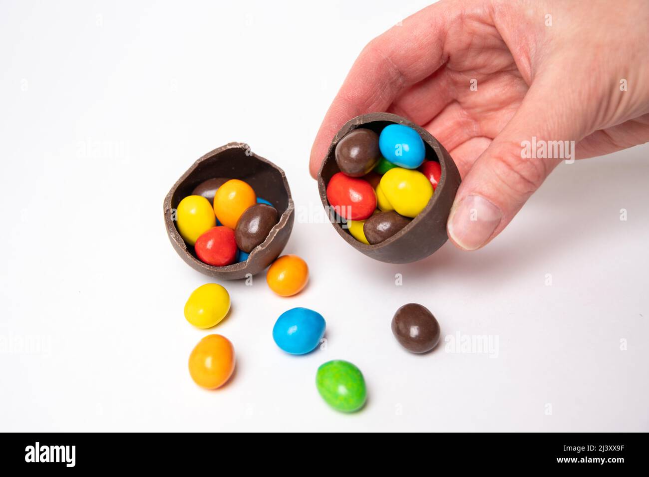 Une main tenant un œuf de pâques au chocolat craqué plein de petites bonbons ronds colorés sur un fond blanc avec des bonbons colorés. Chocolat pour ki Banque D'Images