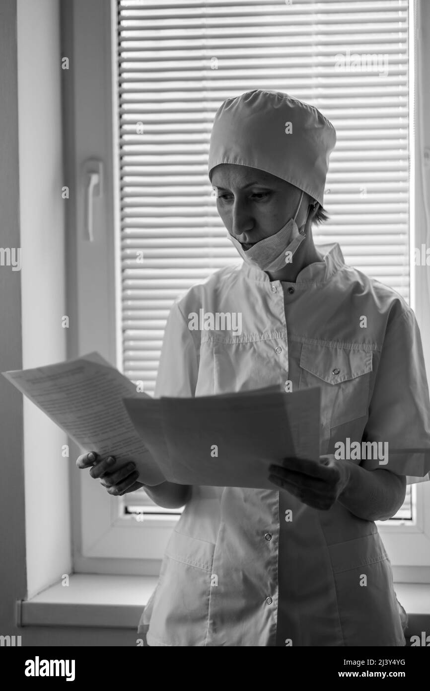 Une femme médecin se tient près de la fenêtre de l'hôpital et regarde à travers les documents. Photo en noir et blanc. Banque D'Images