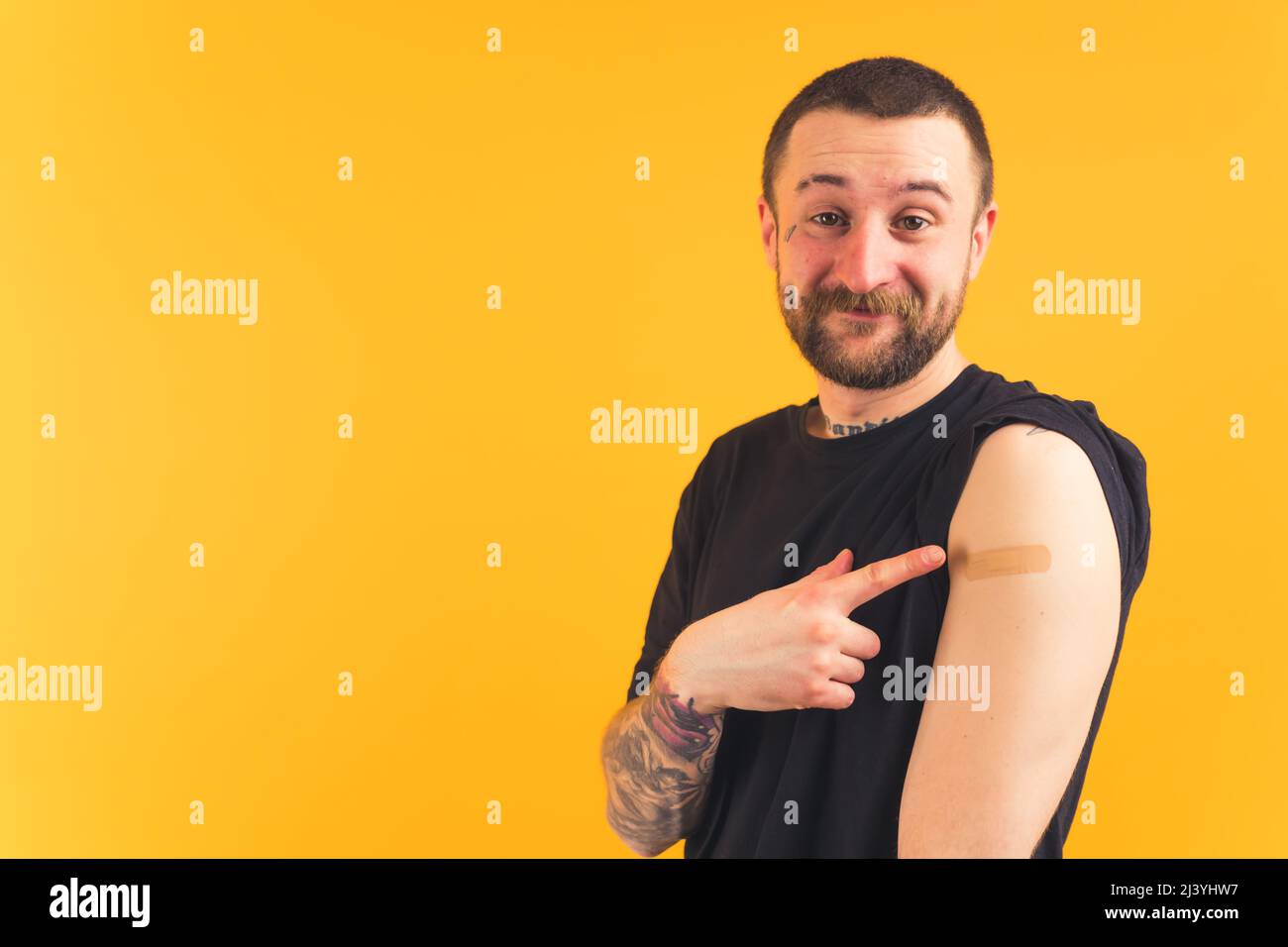 Concept de vaccination. Heureux gangster-comme l'homme caucasien barbu avec des tatouages pointant le doigt vers son bras couvert de patch après la vaccination. Photo de haute qualité Banque D'Images