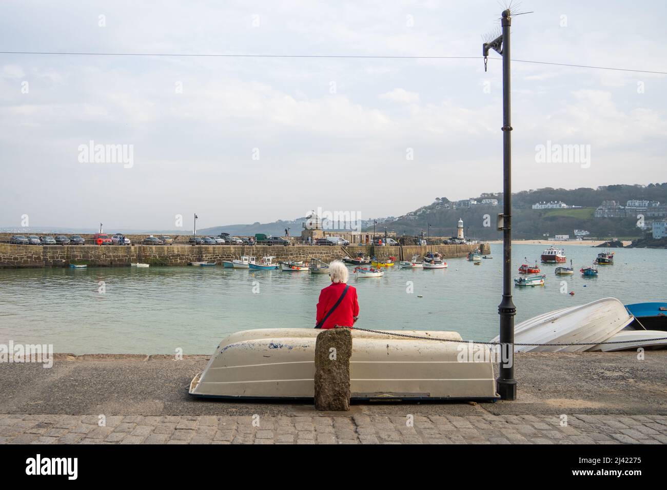 Une femme âgée aux cheveux blancs et au pelage rouge vif se trouve devant un bateau retourné, face à Smeaton's Pier à St Ives, Cornwall, Royaume-Uni. Banque D'Images