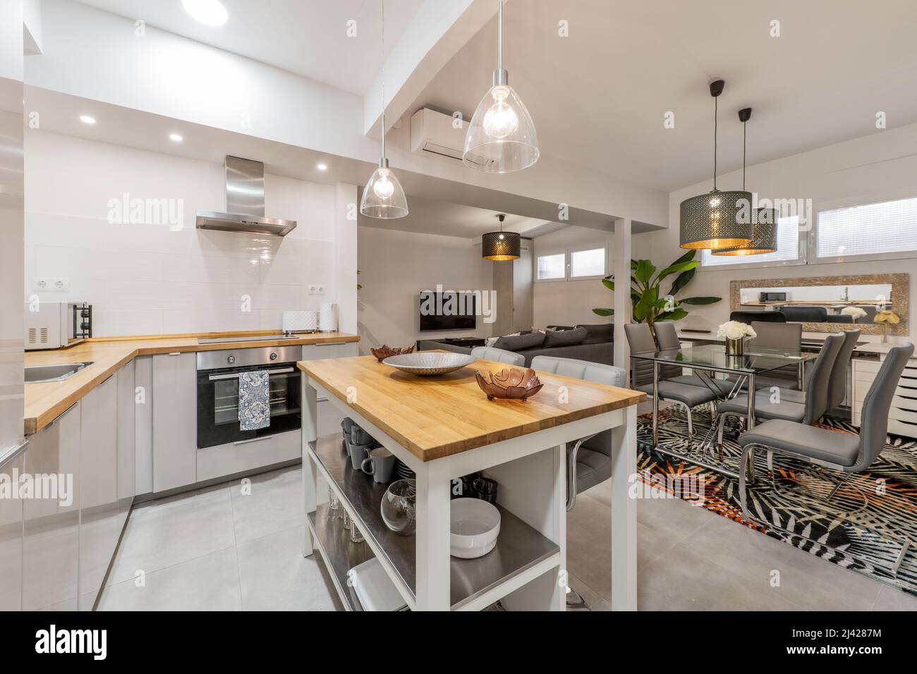 Image d'une cuisine avec des comptoirs en bois massif, des appareils en acier inoxydable, des abat-jour transparents, une île assortie avec de grands chaises grises, et un salon ro Banque D'Images