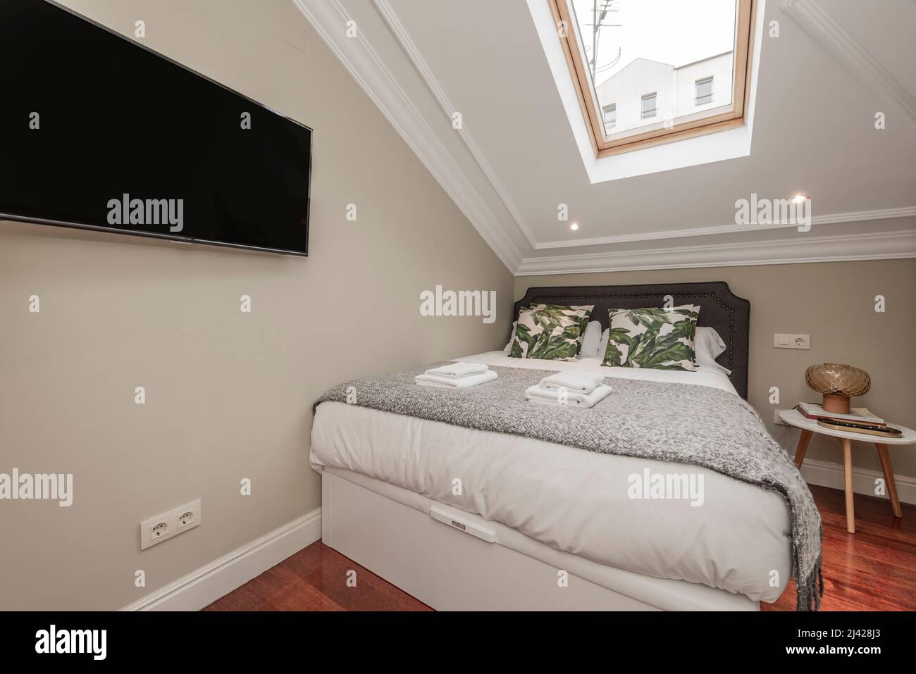 Chambre avec lit double avec coussins et couvertures en denim, coussins verts, lucarne au plafond, tv accrochée au mur, serviettes blanches dans le grenier Banque D'Images