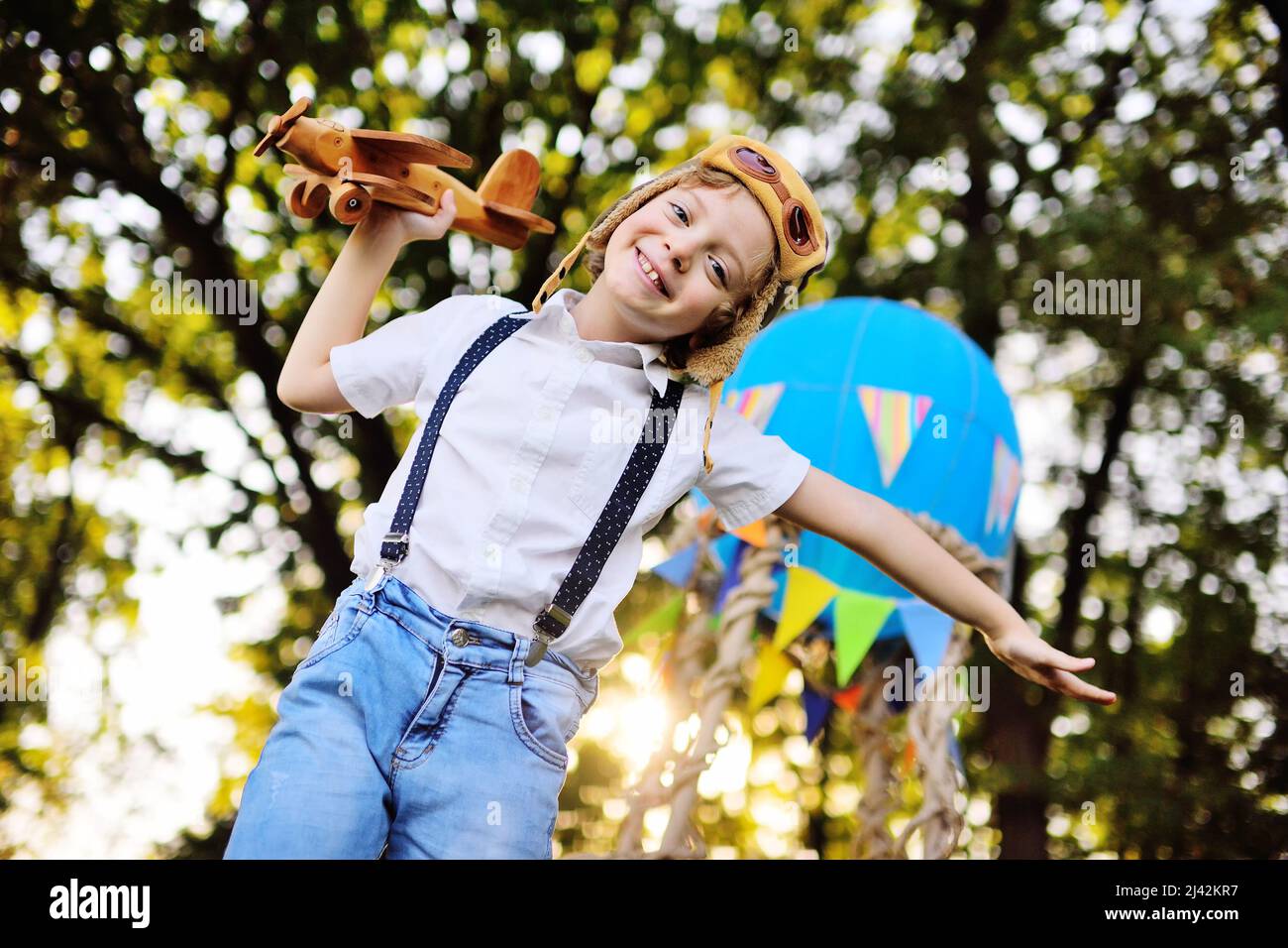 petit garçon dans une chemise blanche avec bretelles et boucles cheveux  dans un chapeau de pilote et des lunettes joue avec un plan en bois contre  le fond d'un panier Photo Stock -