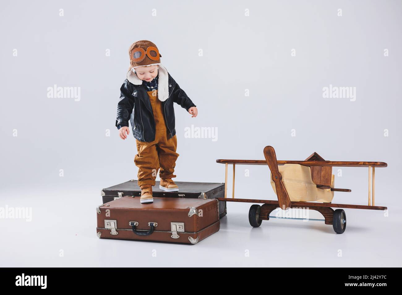 Un garçon heureux dans un casque et une veste de pilote se tient près d'un avion en bois. Portrait d'un pilote d'enfant, d'un enfant dans une veste en cuir. Jouets en bois. Plan ECO Banque D'Images