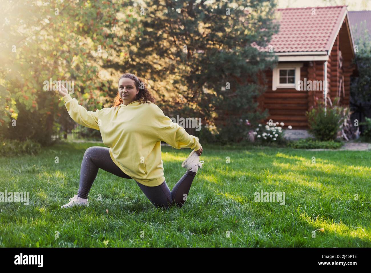 Femme d'âge moyen surpondérée faisant du yoga dans des vêtements pour le sport sur l'herbe verte dans la cour avec maison de campagne en bois et de grands arbres en arrière-plan Banque D'Images
