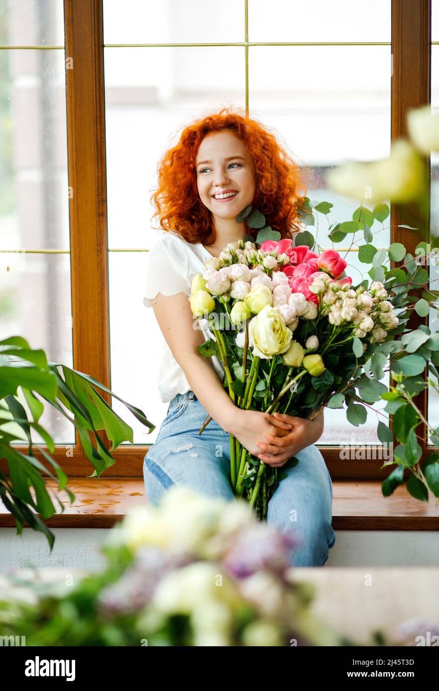 Belle fleuriste femme avec des cheveux rouges bouclés tenant des fleurs fraîchement fait bouquet de fleurs avec des pivoines, roses, eucalyptus sur fond de fenêtre Banque D'Images