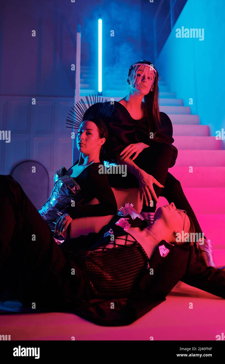 Groupe de trois jeunes artistes de danse vogue en tenue de posh posant sur l'escalier d'un studio moderne de loft éclairé par des néons roses et bleus Banque D'Images