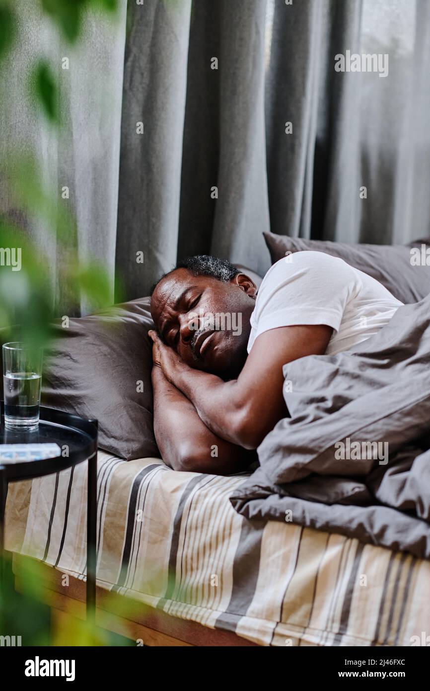 Homme afro-américain mature et fatigué dormant paisiblement au lit le matin devant une petite table noire avec un verre d'eau Banque D'Images