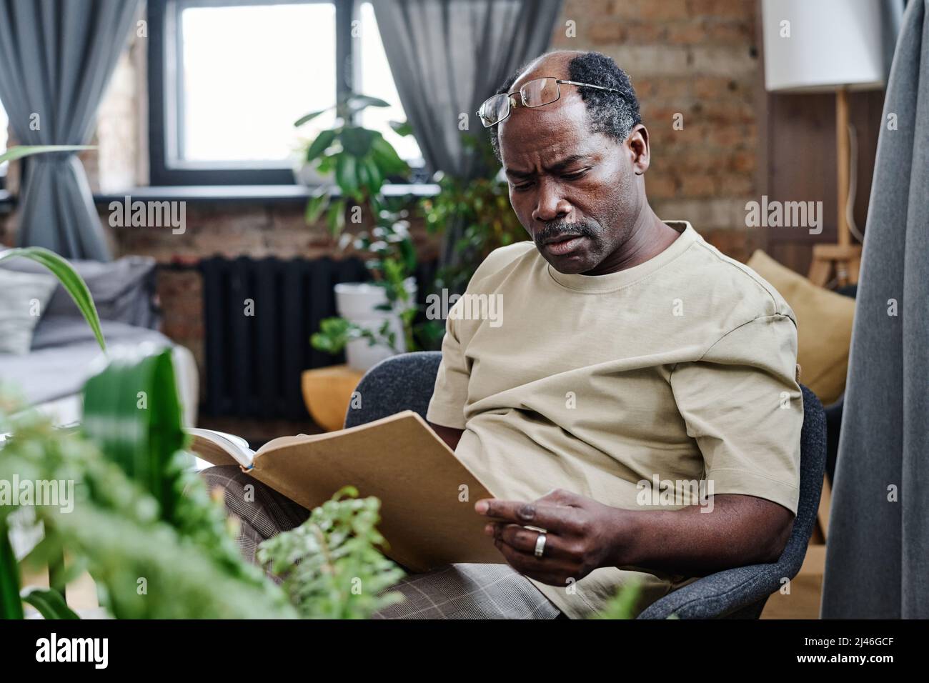 Un homme à la retraite afro-américain contemporain assis dans un fauteuil et lisant un livre curieux à votre guise lors de votre séjour à la maison Banque D'Images