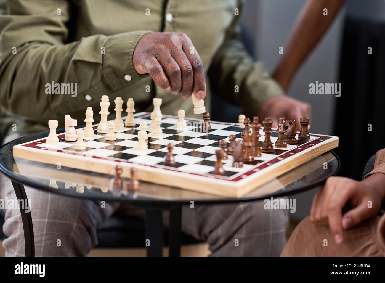 Main de l'homme afro-américain senior qui met une figure blanche sur le plateau d'échecs tout en jouant à un jeu de loisirs avec ses deux petits-enfants Banque D'Images