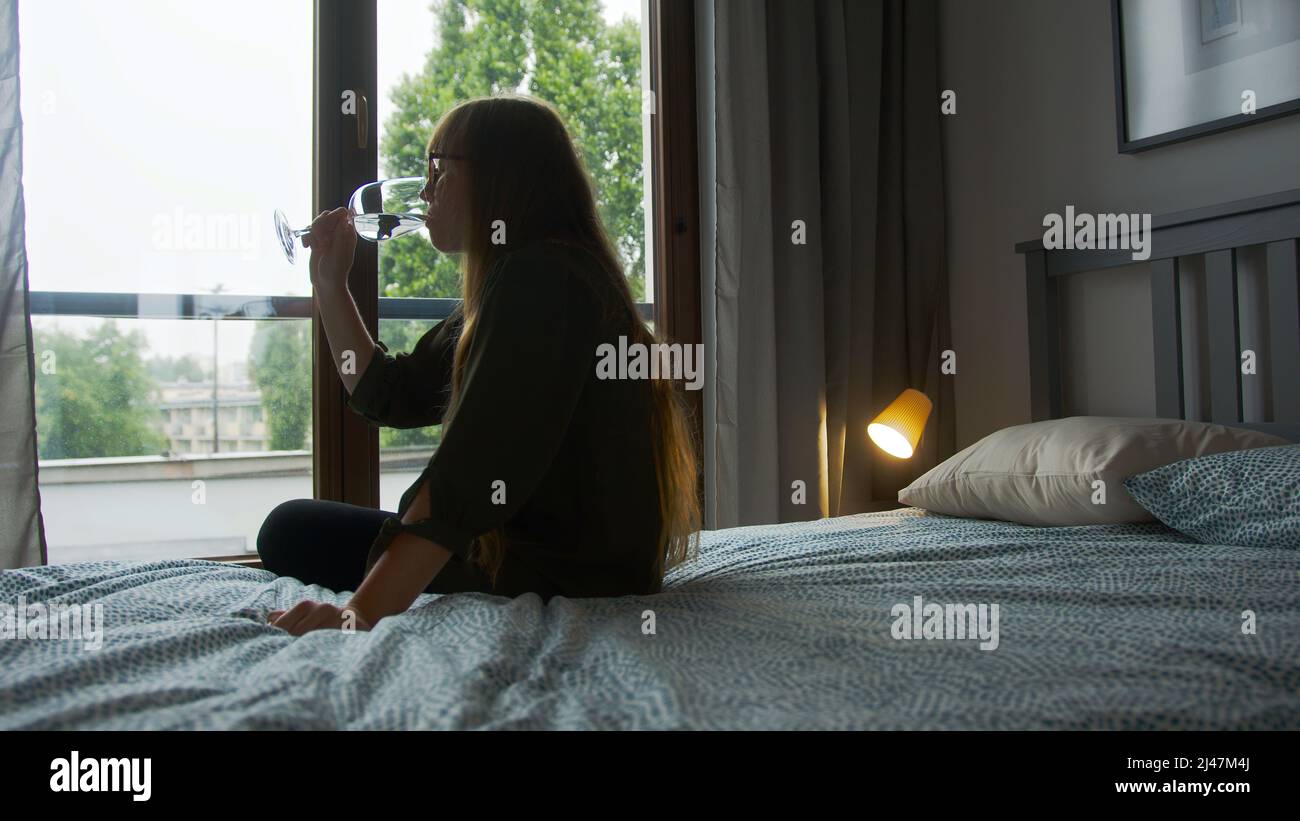 Notion de dépendance à l'alcool. Femme caucasienne solitaire triste à cheveux longs qui boit un autre verre de vin tout en étant assise sur un lit. Photo de haute qualité Banque D'Images