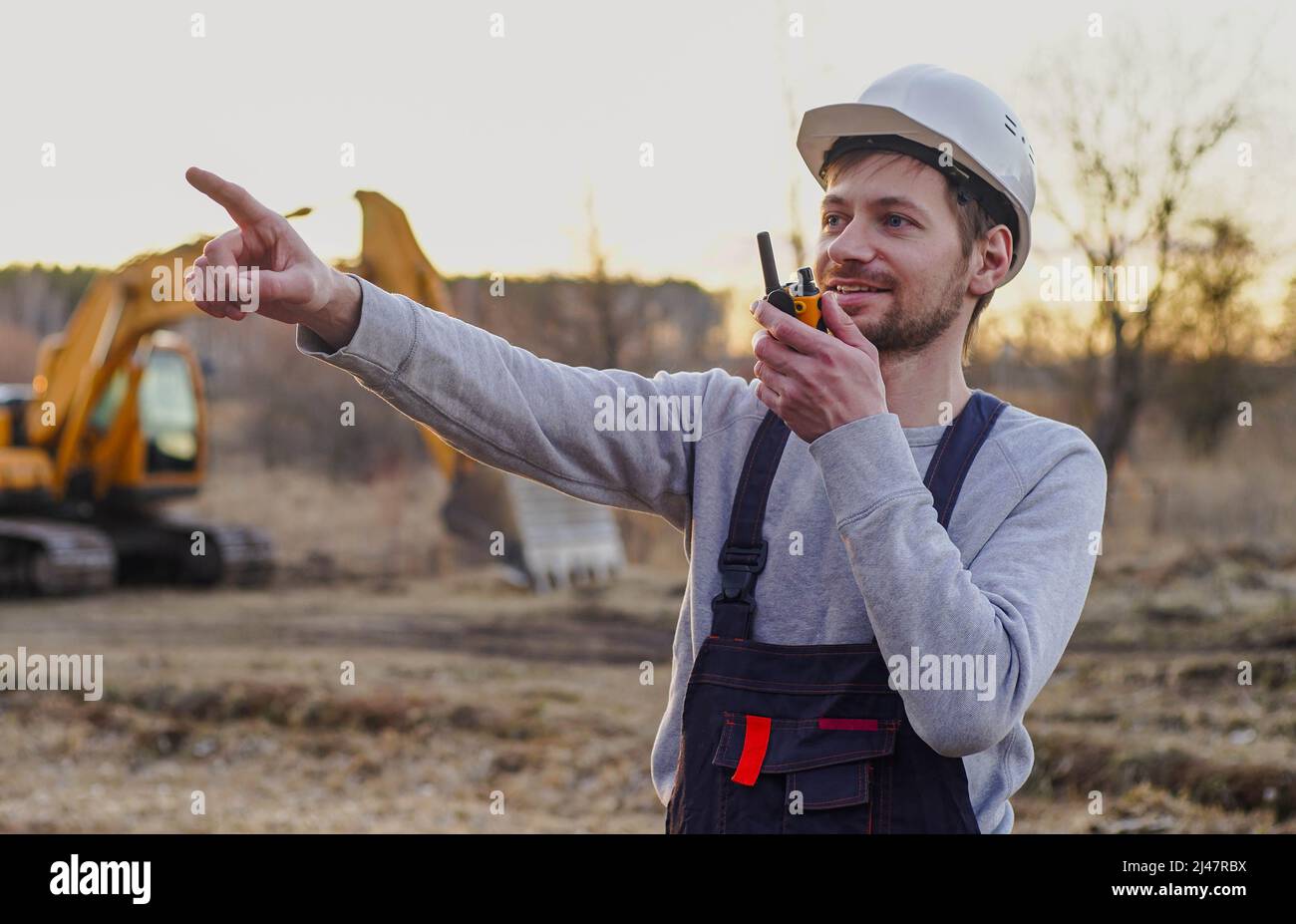 Travailleur dans un casque parlant par émetteur radio sur le chantier de  construction. Pelle hydraulique en arrière-plan Photo Stock - Alamy