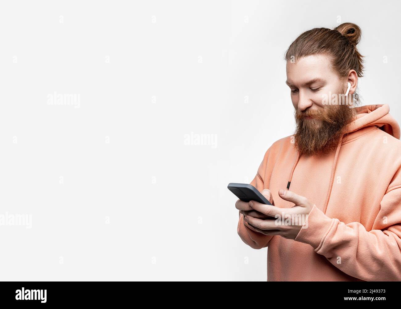 Homme de bonne humeur scandinave tenant le téléphone et souriant avec un casque sans fil dans un sweat-shirt orange isolé sur fond gris. Gars avec gingembre h Banque D'Images
