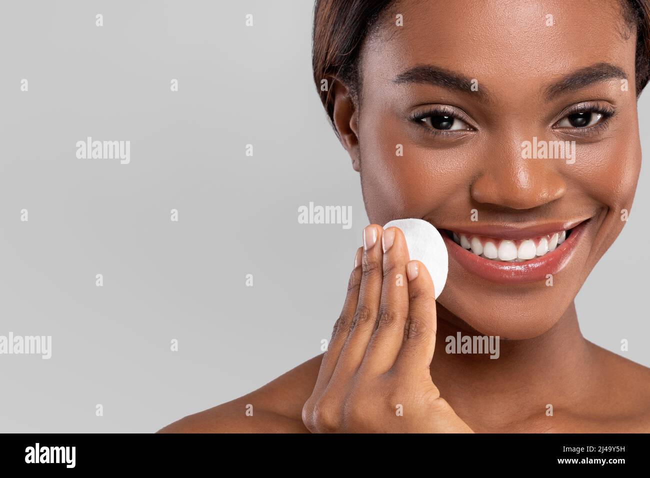 Souriante jolie femme afro-américaine millénaire essuyant son visage avec un tampon en coton, isolé sur fond gris, gros plan Banque D'Images