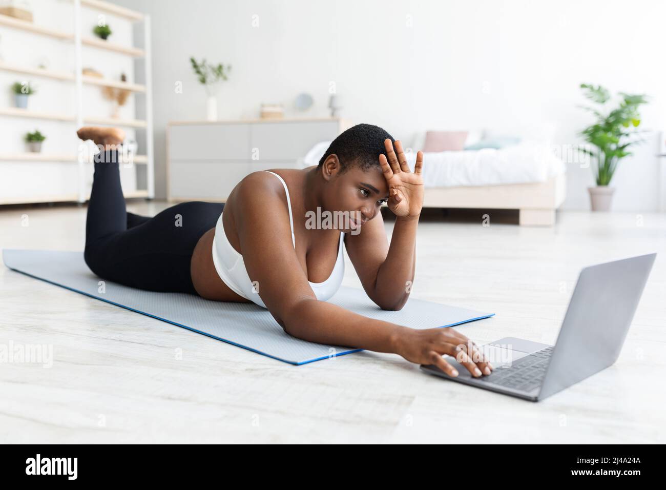 Femme noire fatiguée de plus grande taille, couchée sur un tapis de yoga près d'un ordinateur portable, essuyant le front après un entraînement de force à la maison Banque D'Images