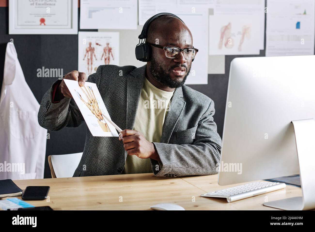 Traumatitologue africain dans les écouteurs pointant sur l'image avec des parties du corps et enseignant en ligne à l'aide de l'ordinateur au bureau Banque D'Images