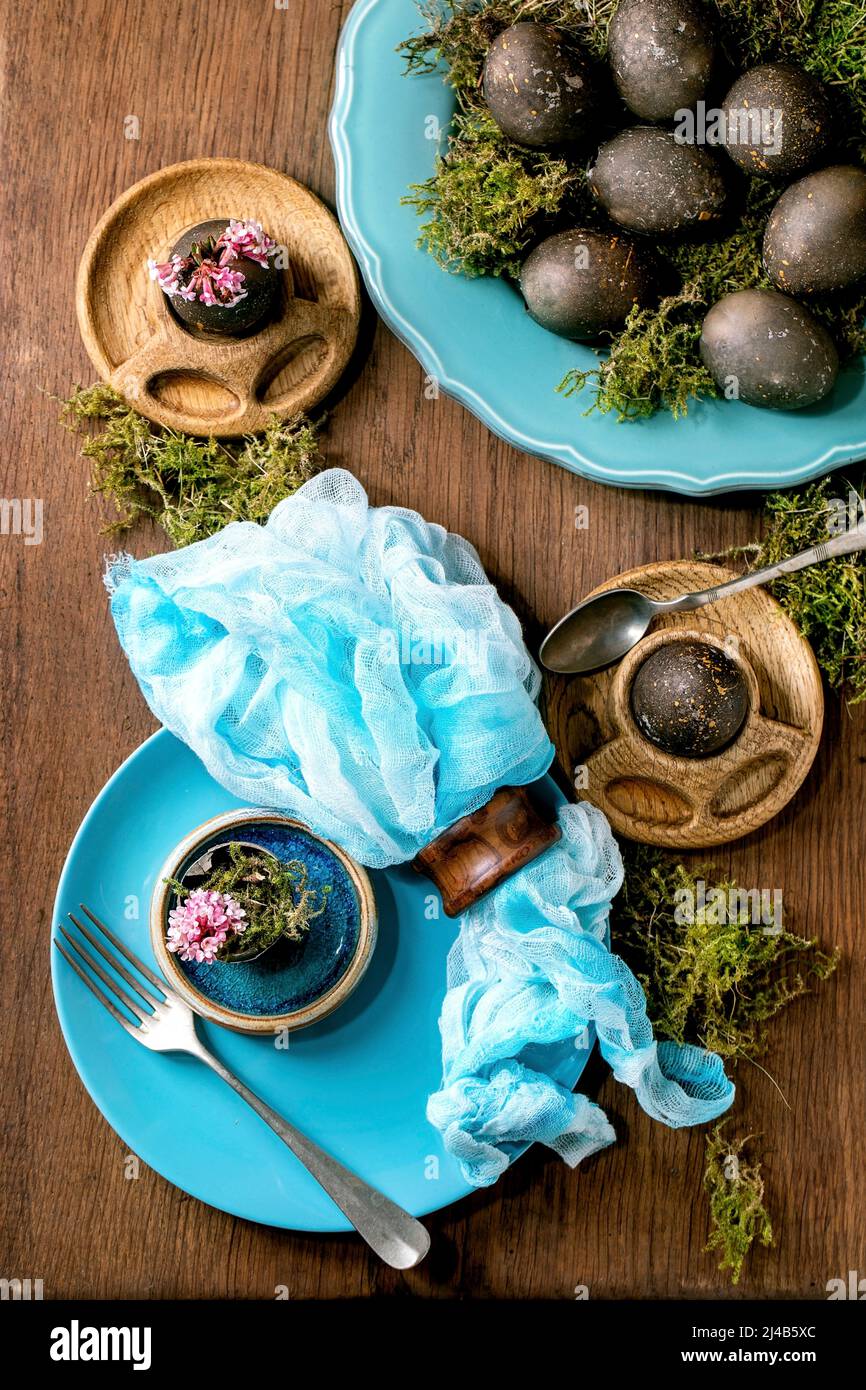 Table de Pâques avec assiettes en céramique turquoise et coquetiers vides, décorée de mousse forestière, œufs de pâques de couleur noire et fleurs de printemps roses Banque D'Images