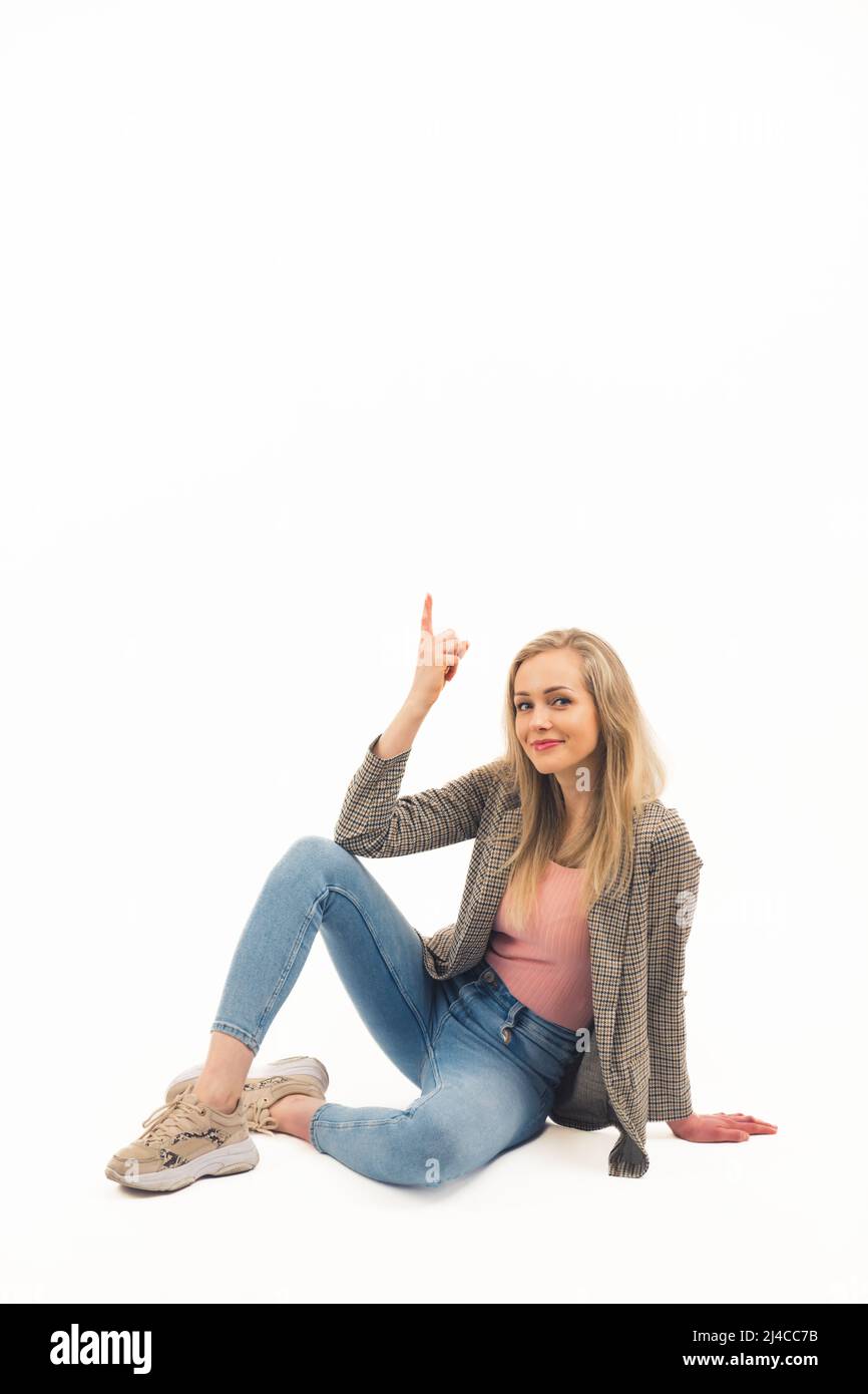 Espace de copie vertical d'une jeune fille blonde vêtue à la mode dans ses 20s en pointant avec son index vers le haut tout en étant assis sur le sol. Arrière-plan blanc. Photo de haute qualité Banque D'Images