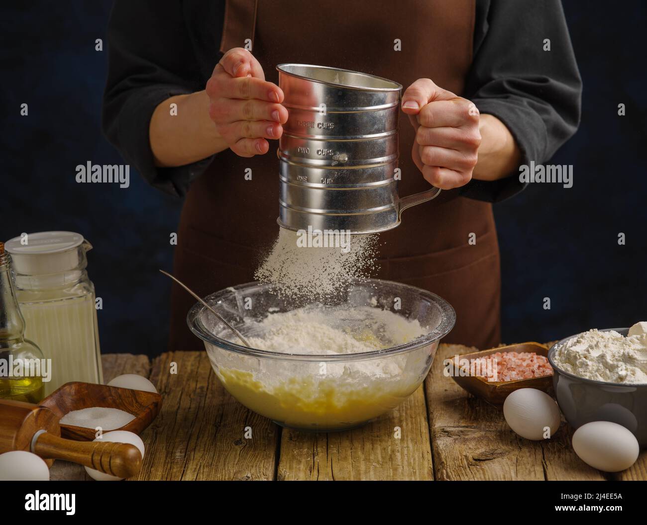 Gros plan. Un chef dans un uniforme sombre est tamisé la farine à travers un tamis sur une table de cuisine en bois. Farine en vol surgelé. Préparation de produits de pâte - Banque D'Images