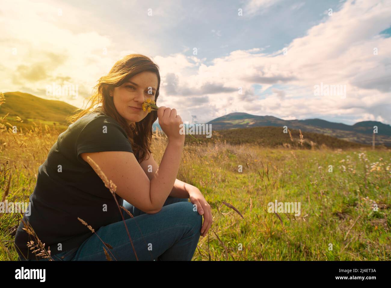 Belle femme hispanique assis regardant l'appareil photo au milieu du champ sentant des fleurs jaunes qu'elle tient dans sa main pendant le soleil Banque D'Images