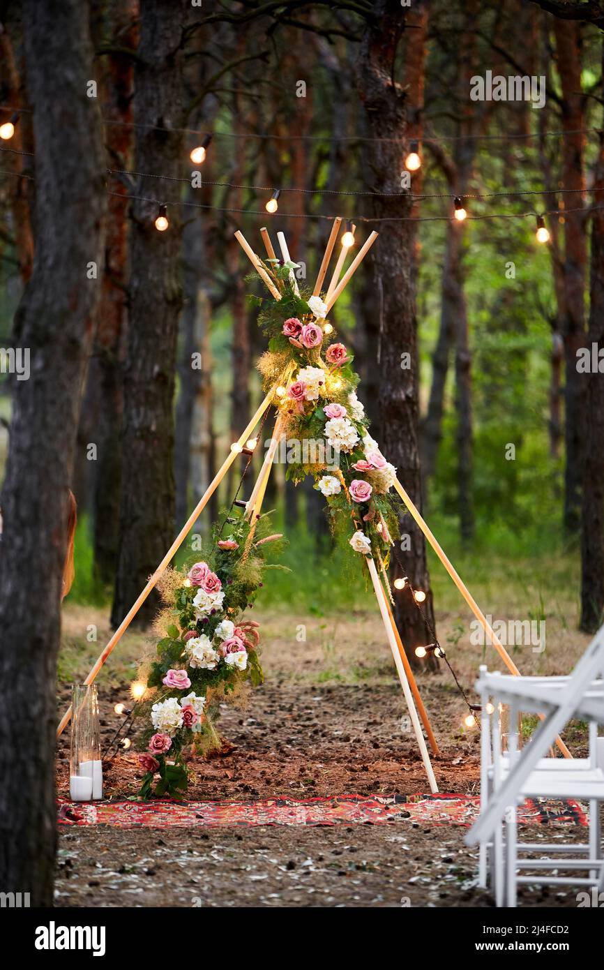 Arche de tipi de Bohème en tiges de bois décorées de roses roses roses,  bougies sur la moquette, herbe de pampass, enveloppée de lumières de fées  sur le mariage extérieur Photo Stock -