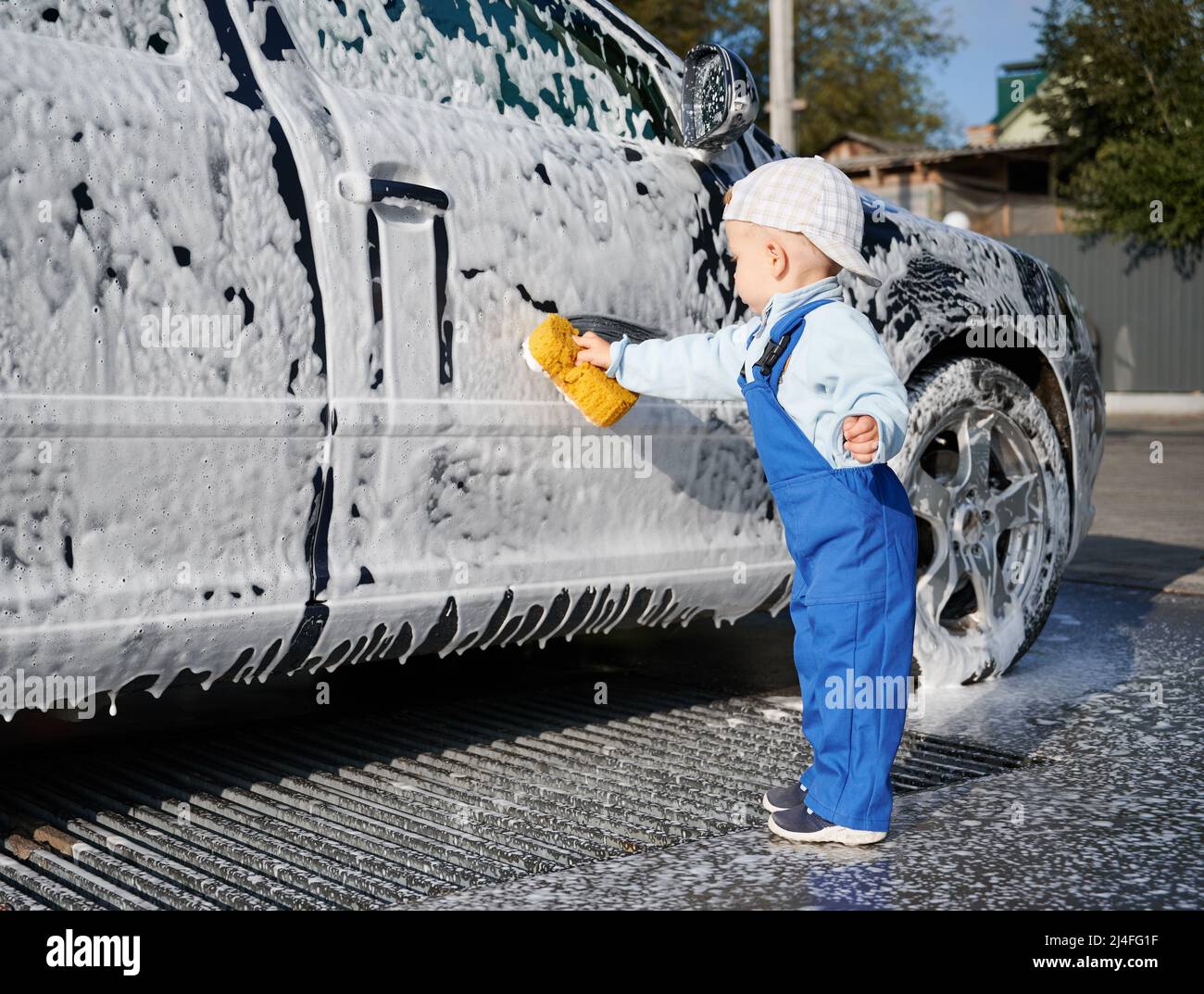 Petit enfant de sexe masculin en combinaison bleue essayant d'étudier comment laver l'automobile, recouvert de mousse blanche à l'aide d'une éponge jaune. Un tout-petit garçon apprend une nouvelle leçon de nettoyage de voiture. Banque D'Images