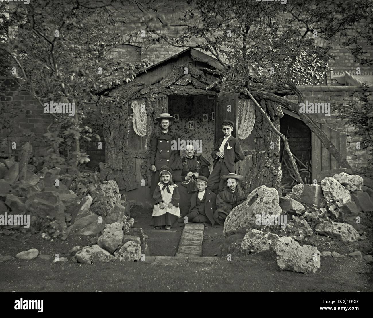 Un groupe familial avec une mère et cinq enfants pose devant leur maison d'été victorienne en Grande-Bretagne vers 1900. La maison d'été rustique dispose de fenêtres avec des rideaux en filet, du papier peint et des images sur le mur. À côté d'elle est un hangar avec l'espace de stockage. Devant la famille se trouve un impressionnant jardin de rochers avec des rochers et des rochers massifs. Ceci est tiré d'un négatif en verre victorien – une photographie vintage 1800s/1900s. Banque D'Images