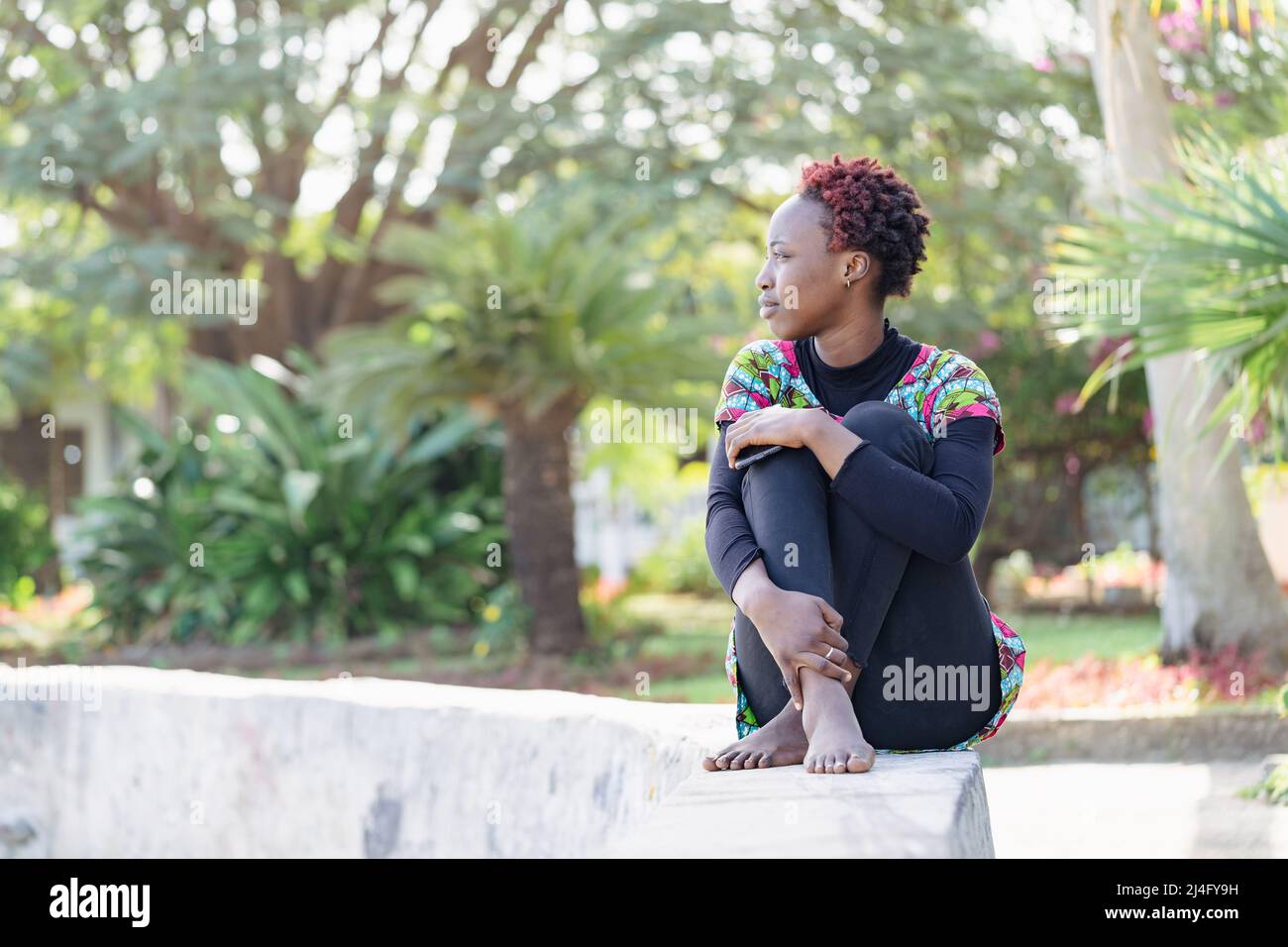 Jeune fille africaine décontractée assise à pieds croisés sur un mur bas dans le parc, attendant que ses amis viennent faire quelque chose ensemble Banque D'Images
