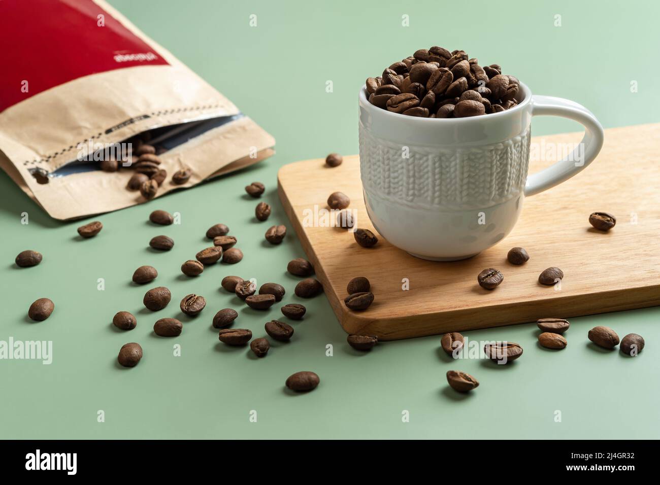 Grains de café torréfiés dans une petite tasse blanche sur fond vert pastel. Grains de café entiers déversés d'un sac de papier brun. Grains de café biologiques. Banque D'Images