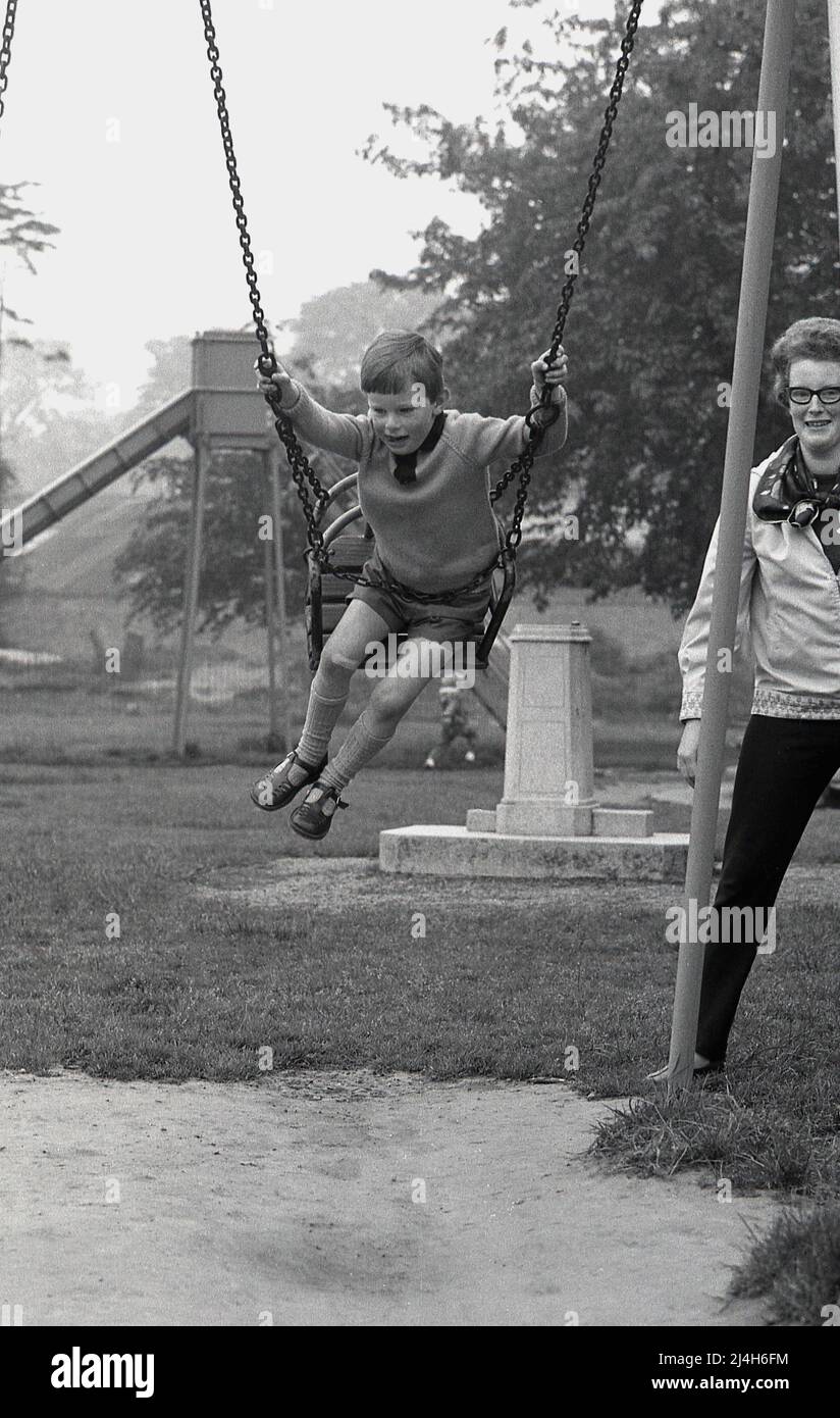 1969, historique, un jeune élève dans un parc sur une balançoire de l'ea, tenant à la chaîne de cordes métalliques, regardé par sa mère, Angleterre, Royaume-Uni. Garçon typique avec un plâtre sur son genou et portant des sandales sur ses pieds, des chaussures communes pour les jeunes à ce moment. Banque D'Images