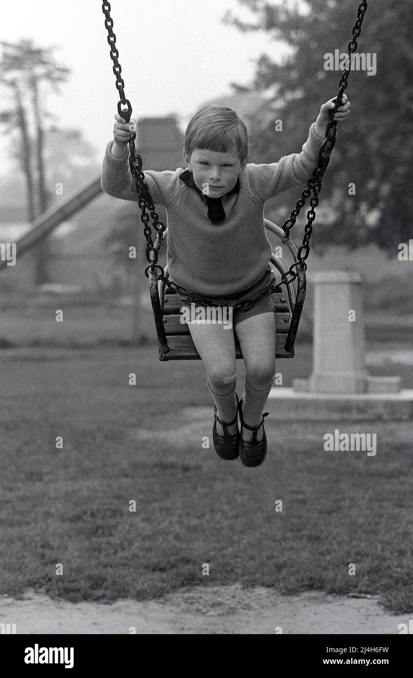1969, historique, un jeune garçon dans un parc sur un terrain de jeu balançoire de l'époque, assis sur la chaise en bois et tenant sur les chaînes métalliques, Angleterre, Royaume-Uni. Garçon typique avec un plâtre sur son genou et portant des sandales sur ses pieds, des chaussures communes pour les jeunes à ce moment. Banque D'Images