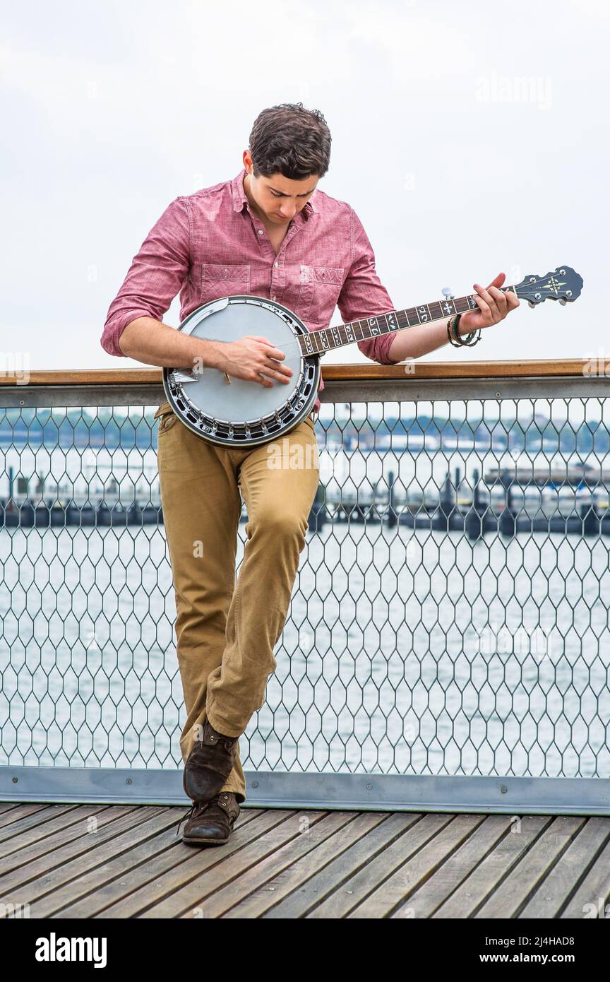 Avec sa chemise rouge clair, son pantalon jaune foncé et ses chaussures brunes, un jeune musicien se pose sur le banjo Banque D'Images
