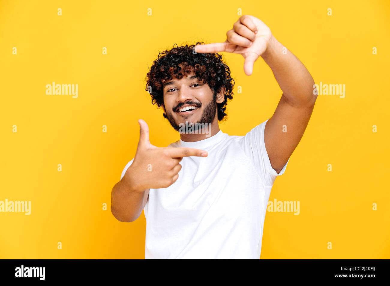 Joyeux joyeux joyeux garçon millénaire indien ou arabe en t-shirt blanc de base, faisant cadre imaginaire de l'appareil photo prise de vue, debout sur un arrière-plan orange isolé, regarde l'appareil photo, souriant Banque D'Images