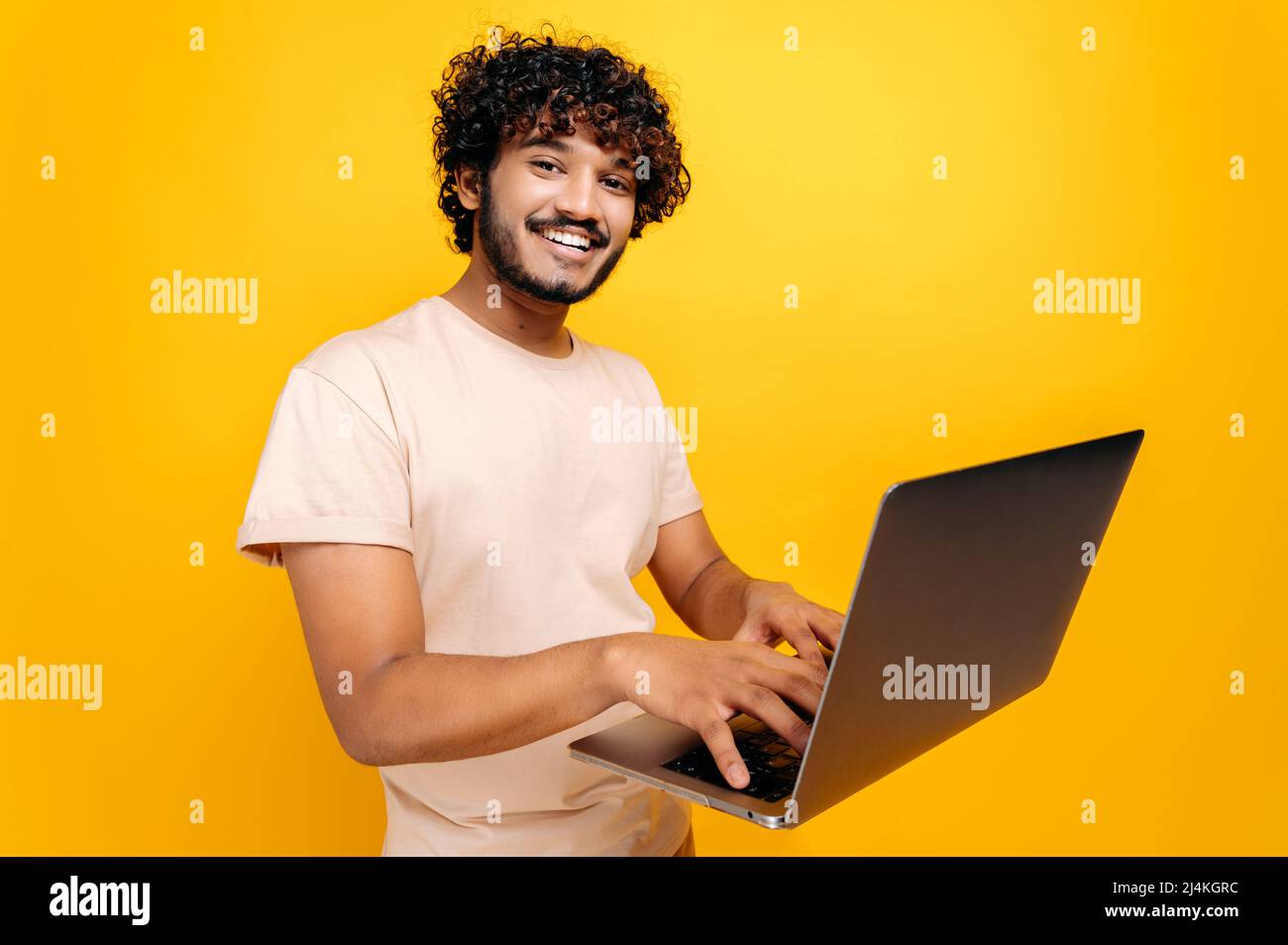 Un homme indien ou arabe millénaires aux cheveux maurires positifs dans un t-shirt, dactylographiant sur un clavier d'ordinateur portable, se tenant sur un arrière-plan orange isolé, regardant l'appareil photo, souriant amical. Concept de technologie Banque D'Images