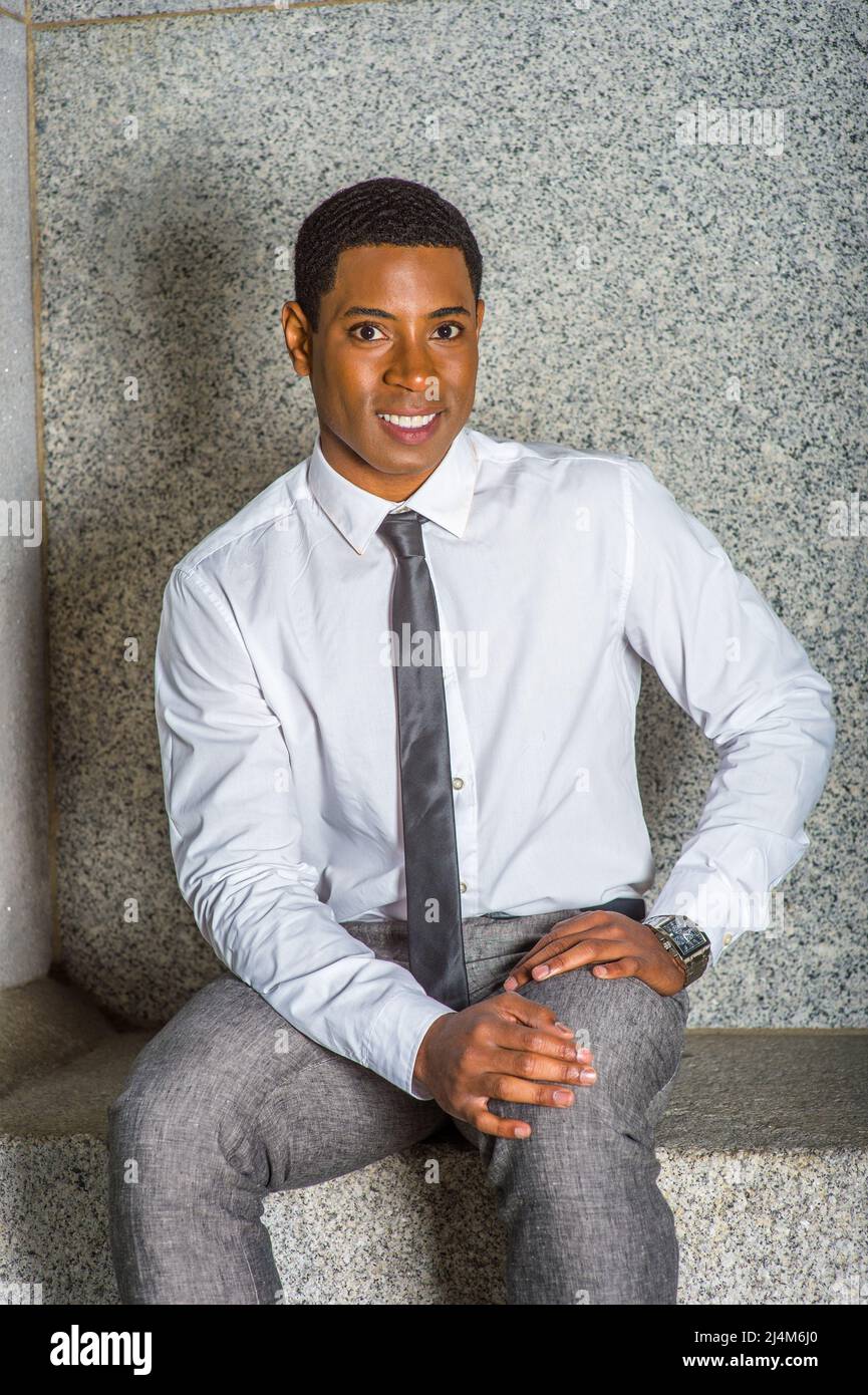 Dressing en chemise blanche, une cravate noire, pantalon gris, mains  reposant sur ses genoux, un jeune beau étudiant noir d'université est assis  sur un banc de pierre, smilin Photo Stock - Alamy