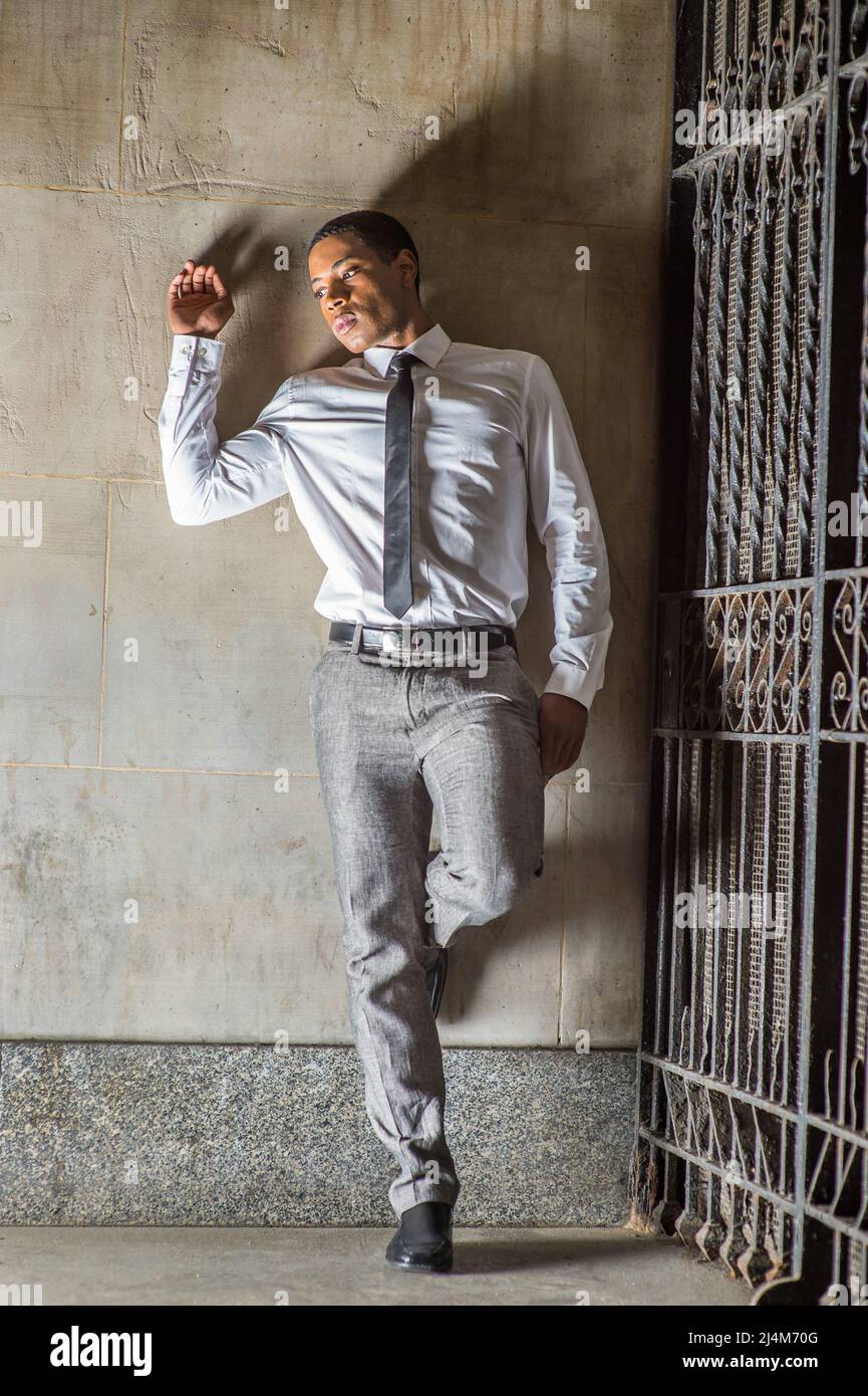 Portant une chemise blanche une cravate noire un pantalon gris des chaussures en cuir un jeune homme noir elegant est debout contre un mur a l exterieur d une porte profondement pensant Photo Stock