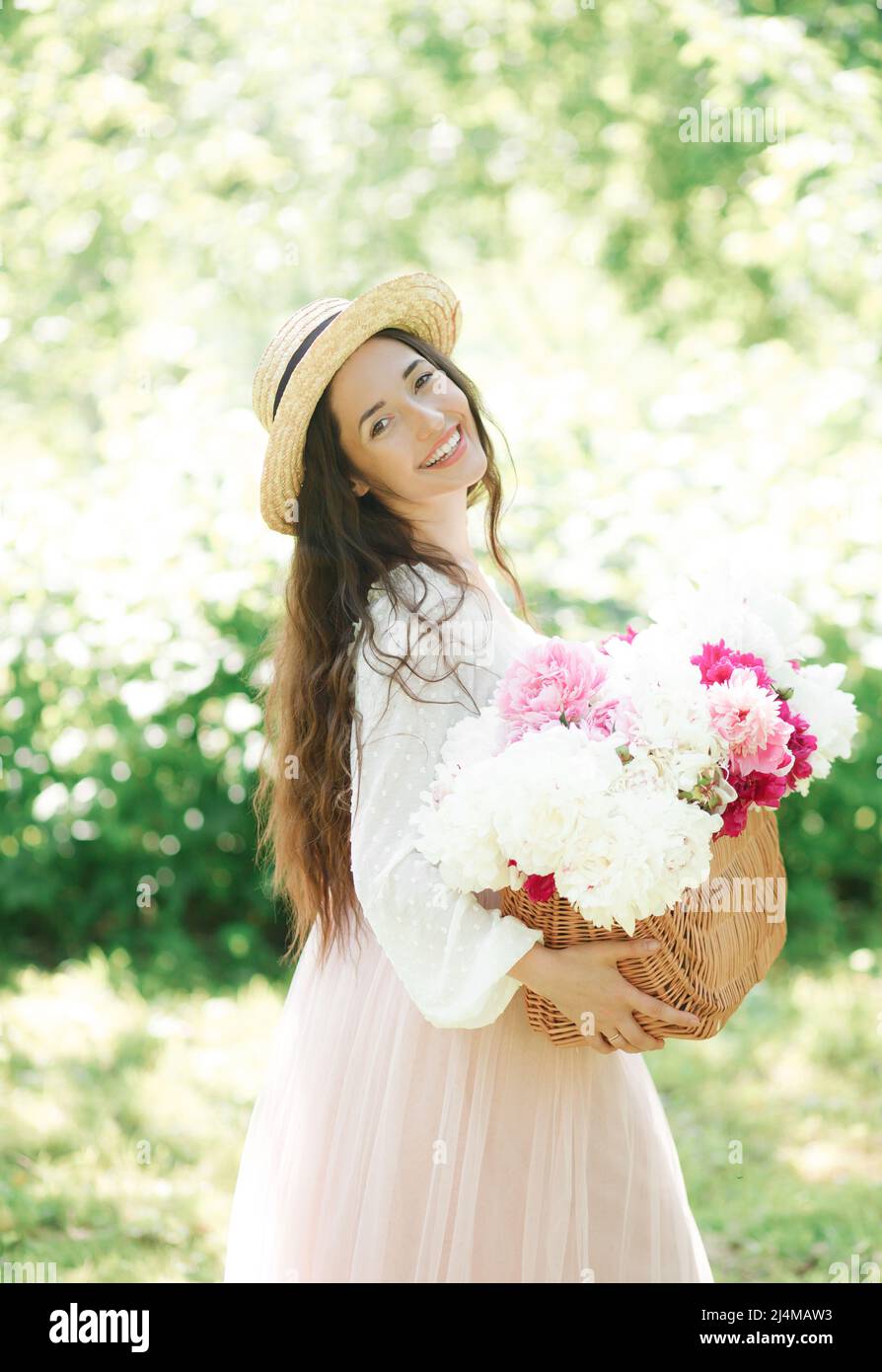 Bonne fille souriante dans un chapeau de paille portant un panier en osier avec des fleurs de pivoine blanches et roses qui se posent dans les rues d'une ville européenne. Style de vie Banque D'Images