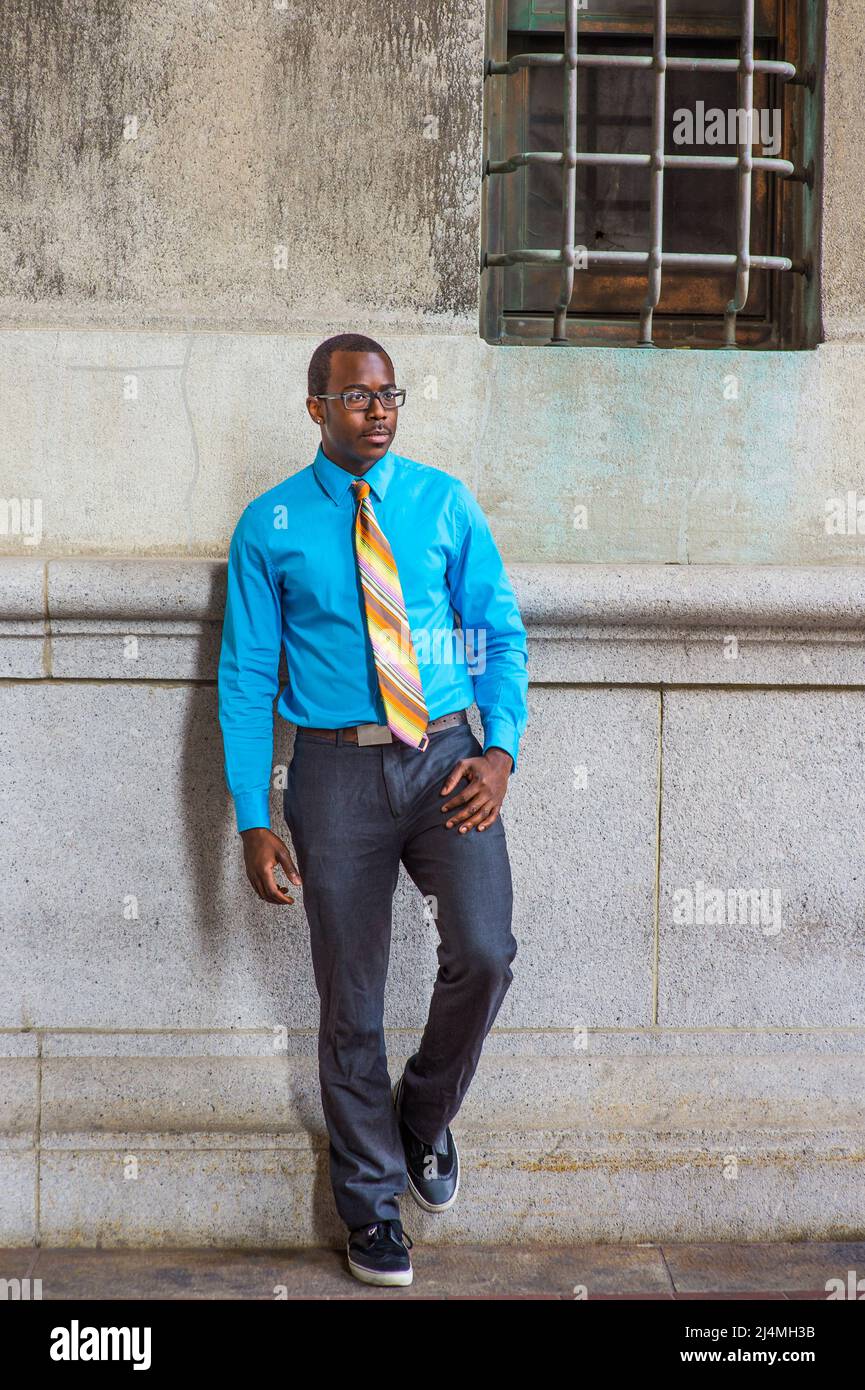 Vêtu d'une chemise bleu clair, d'une cravate à motif, d'un pantalon gris,  portant des lunettes, un étudiant d'université noir est debout près d'un  mur à motif avec une fenêtre, confi Photo Stock -