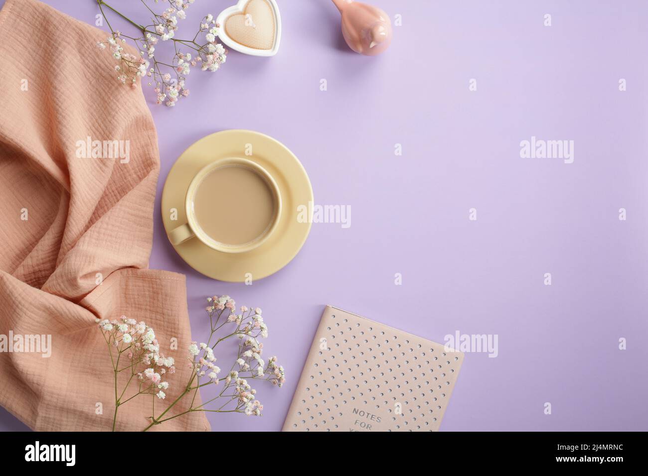 Café du matin, bougie en forme de coeur, carnet rose, foulard beige, décoration et fleurs printanières sur table mauve pastel. Le petit déjeuner romantique est encore vivant. Plat Banque D'Images