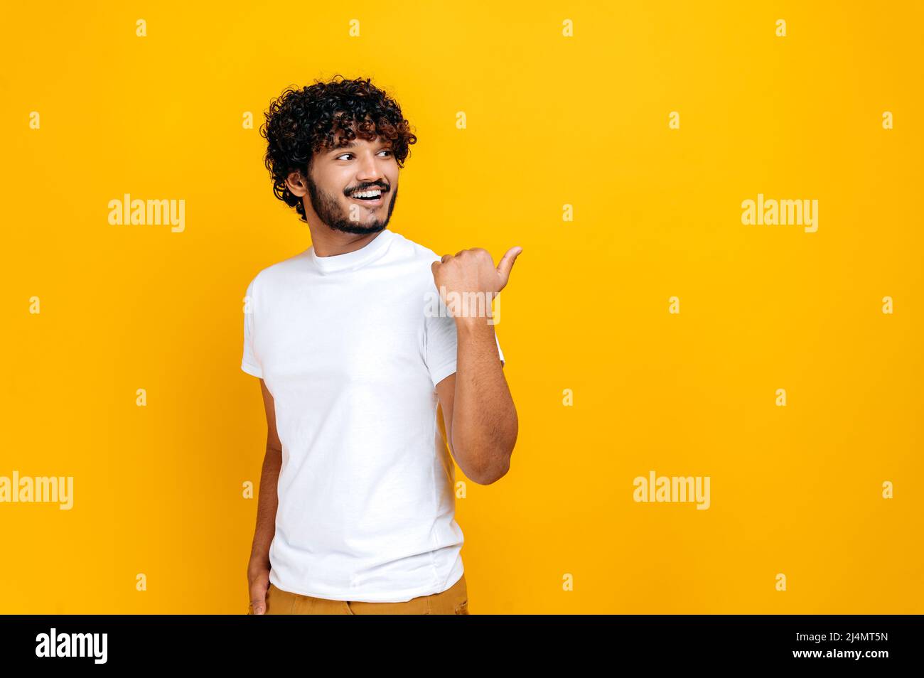 Un homme millénaire indien ou arabe aux cheveux bouclés, regardant et pointant du doigt sur le côté d'un espace de maquette vide, tout en se tenant sur un arrière-plan orange isolé, souriant avec joie Banque D'Images