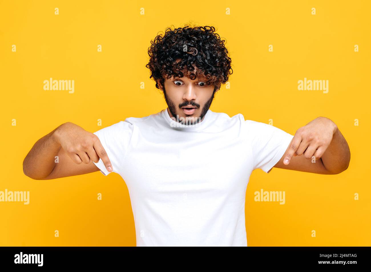 Surprend un indien ou un arabe, un homme aux cheveux bouclés dans un  t-shirt blanc de base, un regard émerveillé et des doigts vers le bas, une  expression du visage excitée, se