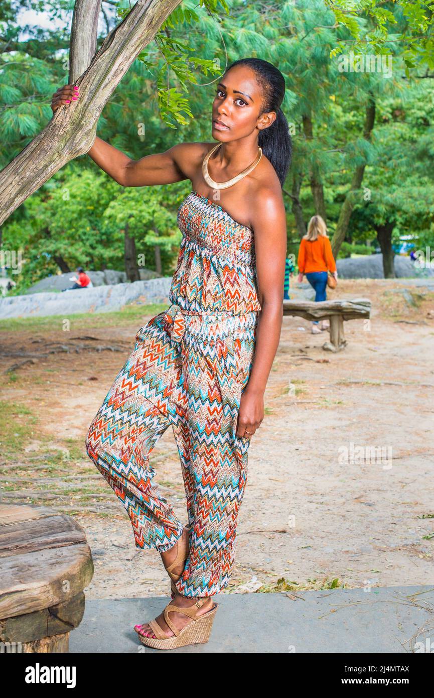 Vêtue d'un haut et d'un pantalon sans bretelles tendance de style contemporain, cette jolie femme noire est debout près d'un arbre dans un parc Banque D'Images