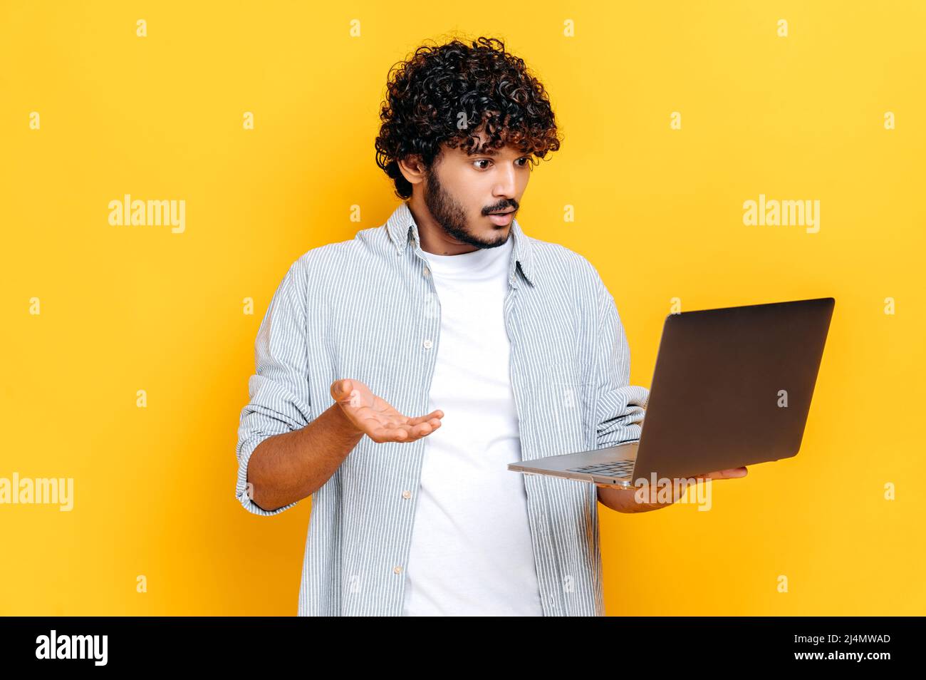 Découragé, un homme millénaire indien ou arabe confus dans des vêtements décontractés, tient un ordinateur portable ouvert à la main, semble perplexe, la main gestante, se tient sur un arrière-plan orange isolé Banque D'Images