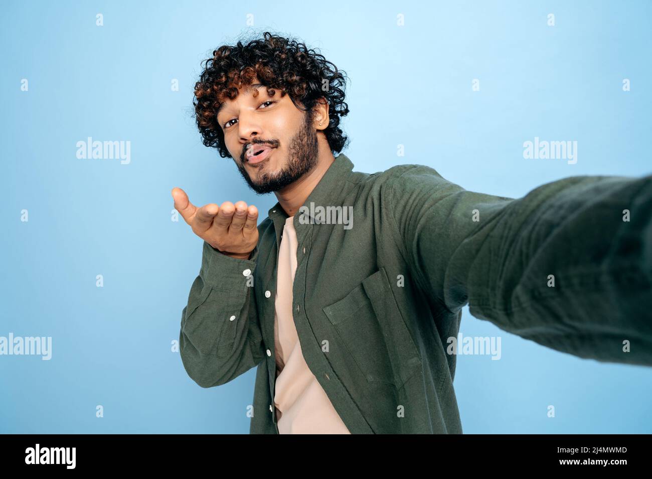 Joyeux Indien positif ou arabe gars excités avec, en tenue décontractée, faisant selfie tourné sur le téléphone portable et sourires dans l'appareil photo, fait un baiser d'air, se tient sur un arrière-plan bleu isolé Banque D'Images