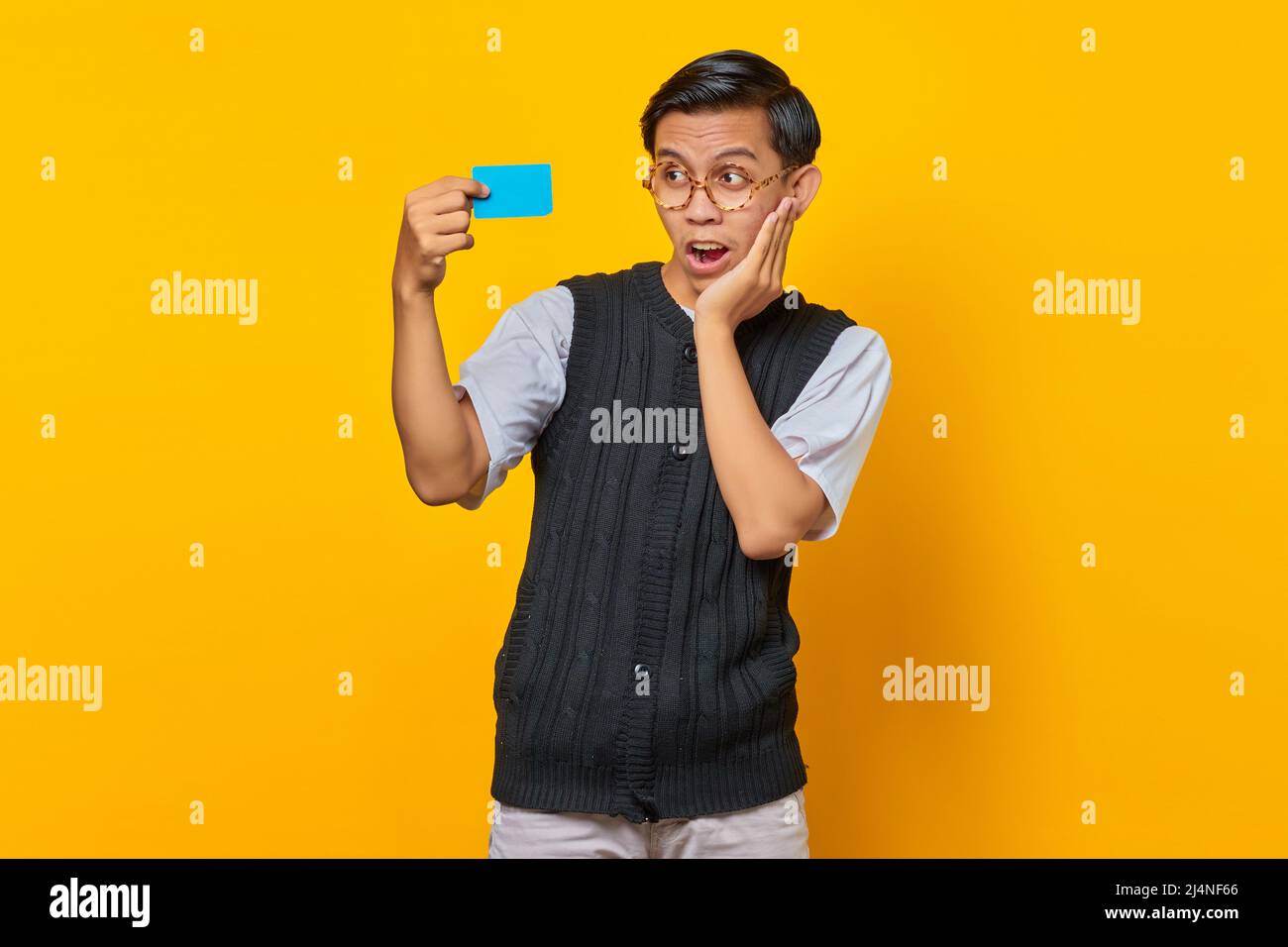 Homme asiatique choqué tenant la carte de crédit avec la bouche ouverte et la main sur une joue sur fond jaune Banque D'Images