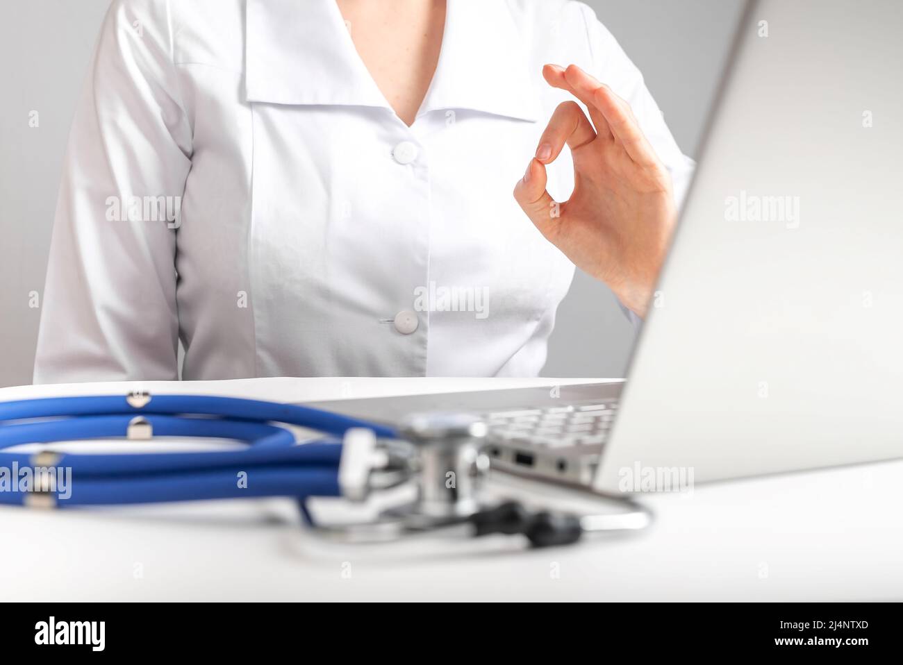 Médecin donnant la consultation en ligne au patient et montrant le geste ok. Télémédecine, concept de télésanté. Femme en blouse de laboratoire assise au bureau avec ordinateur portable et stéthoscope. Photo de haute qualité Banque D'Images