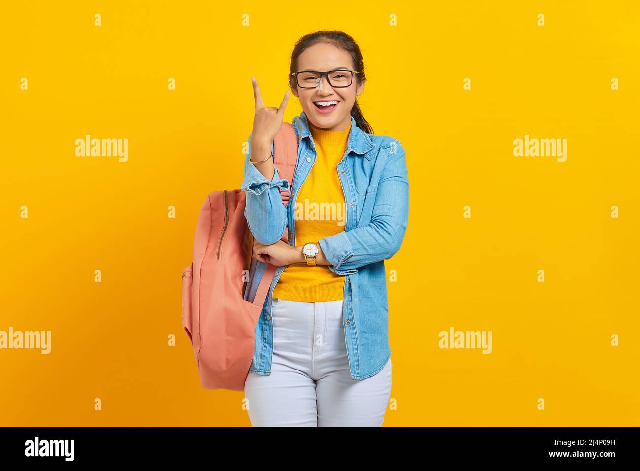 Belle jeune femme asiatique étudiant en tenue denim avec sac à dos criant avec l'expression folle faisant le symbole rock avec les mains isolées sur la bande jaune Banque D'Images