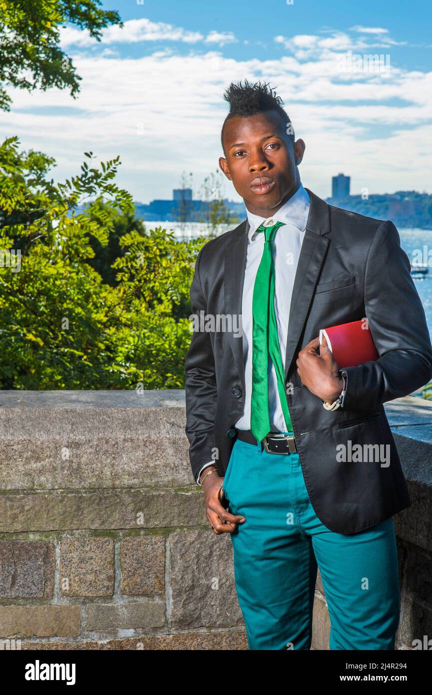 Homme d'affaires moderne. Portant un blazer noir, un pantalon vert, une cravate  verte, une main tenant un livre rouge sous le bras, un jeune homme noir  avec une coupe de cheveux mohawk