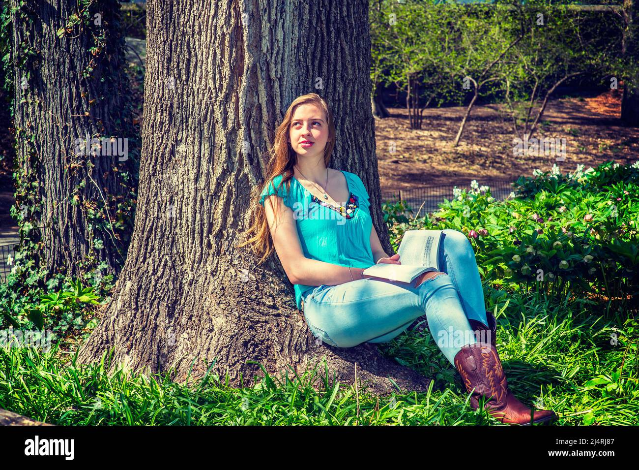 Adolescente américaine portant un haut sans manches bleu, un Jean tendance, des bottes brunes, assis contre le tronc d'arbre à Central Park, New York, lisant rouge b Banque D'Images