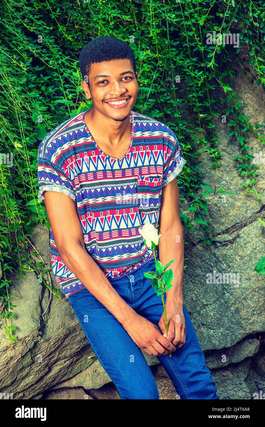 Homme qui vous attend. Vêtu d'une chemise à manches courtes, sans col, à  motif coloré, d'un Jean bleu, un jeune homme noir est assis contre les  rochers avec du vert lea Photo